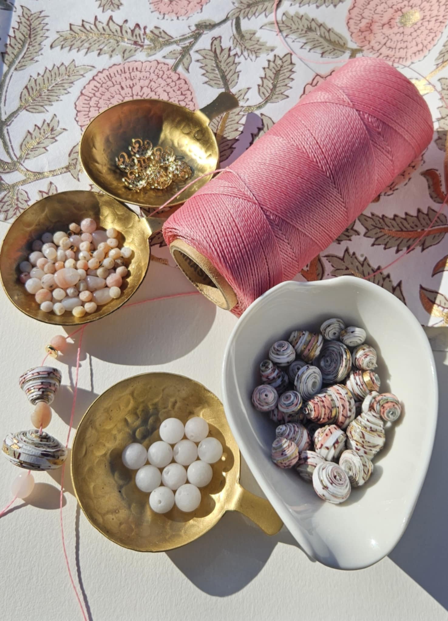 Decorative scene with pink thread, small bowls with beads and shells on a floral background.