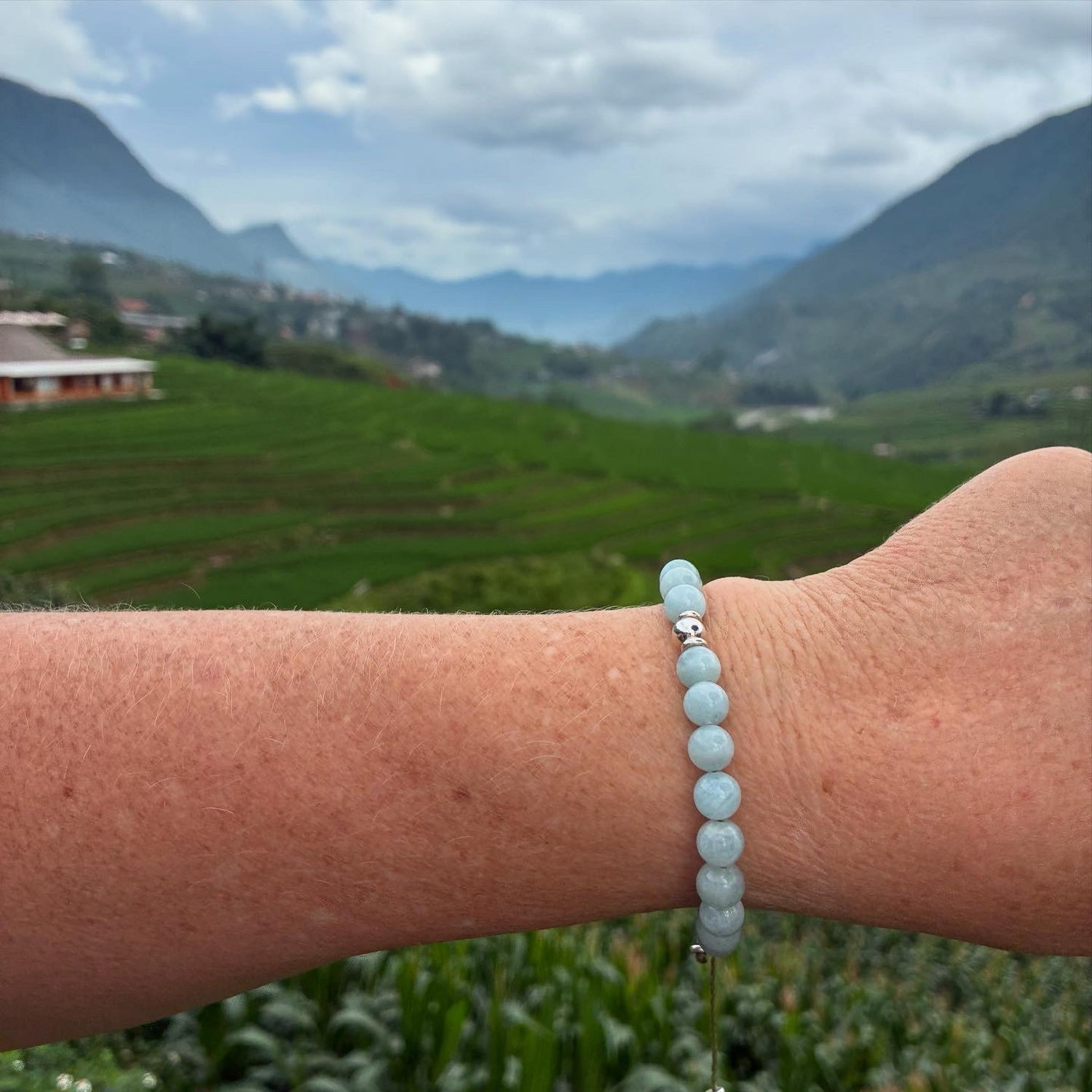 Person wearing a light blue beaded gemstone bracelet with a scenic background of green fields and mountains.