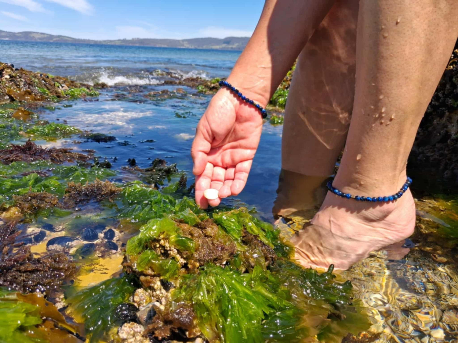 Person sitting in a coastal area with water and rocks in the background wearing a Soul & Stone bracelet and anklet.