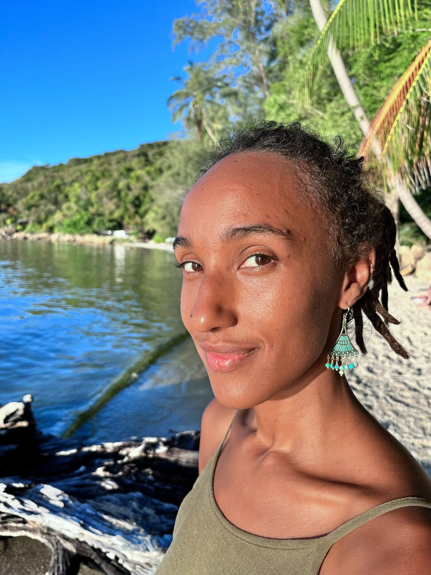 Woman wearing a pair of gemstone earrings standing by a body of water with trees in the background.